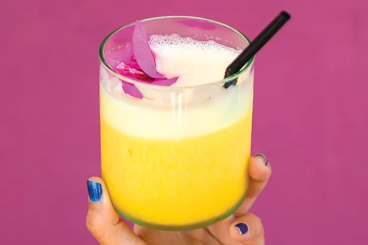 A hand holds a yellow drink with a purple flower garnish against pink.