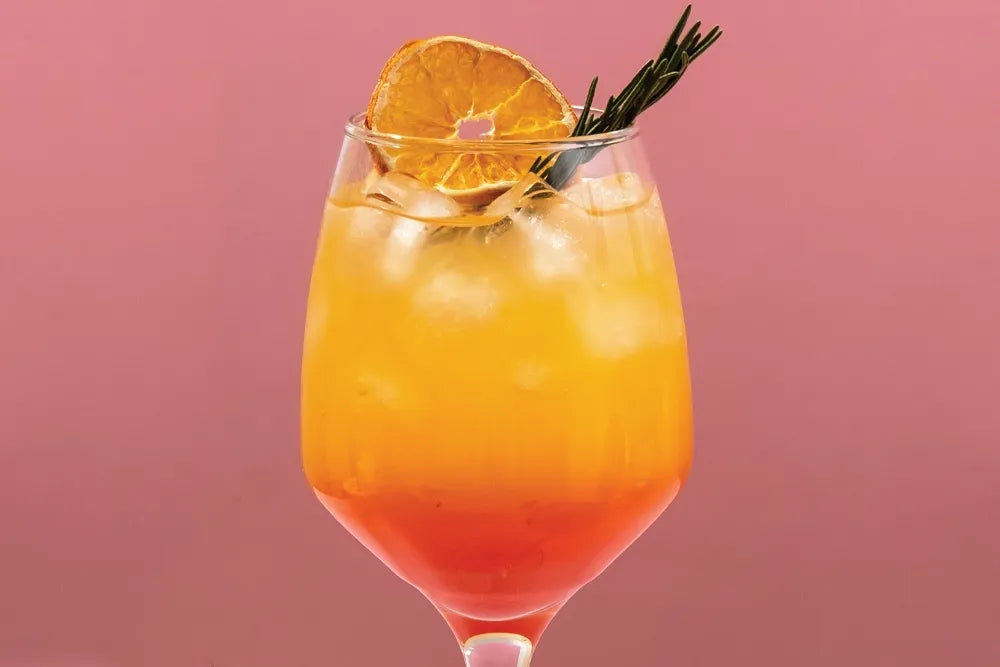 A yellow-and-red cocktail garnished with citrus and rosemary against pink.
