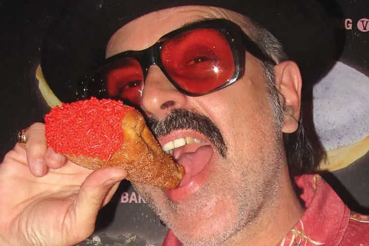Story artwork: A close crop of a person in red glasses taking a bite of a bright red-topped doughnut.