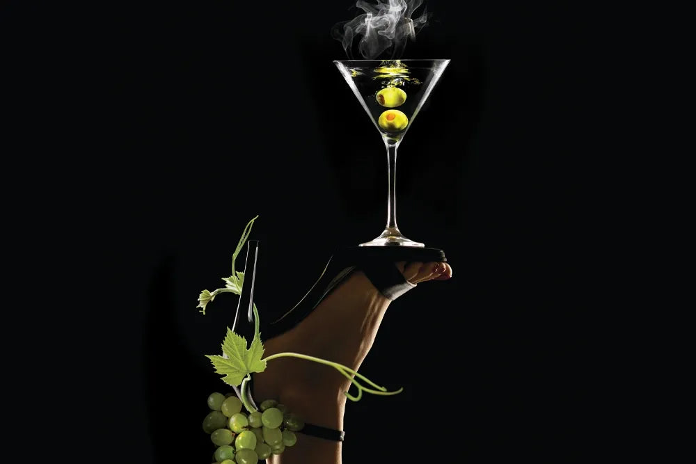 A smoking martini with olives rests on a high-heeled shoe against a black background.