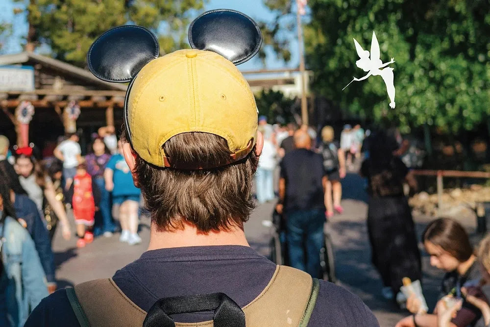 A visitor wearing a yellow Mickey-ear cap looks into a crowded theme park.