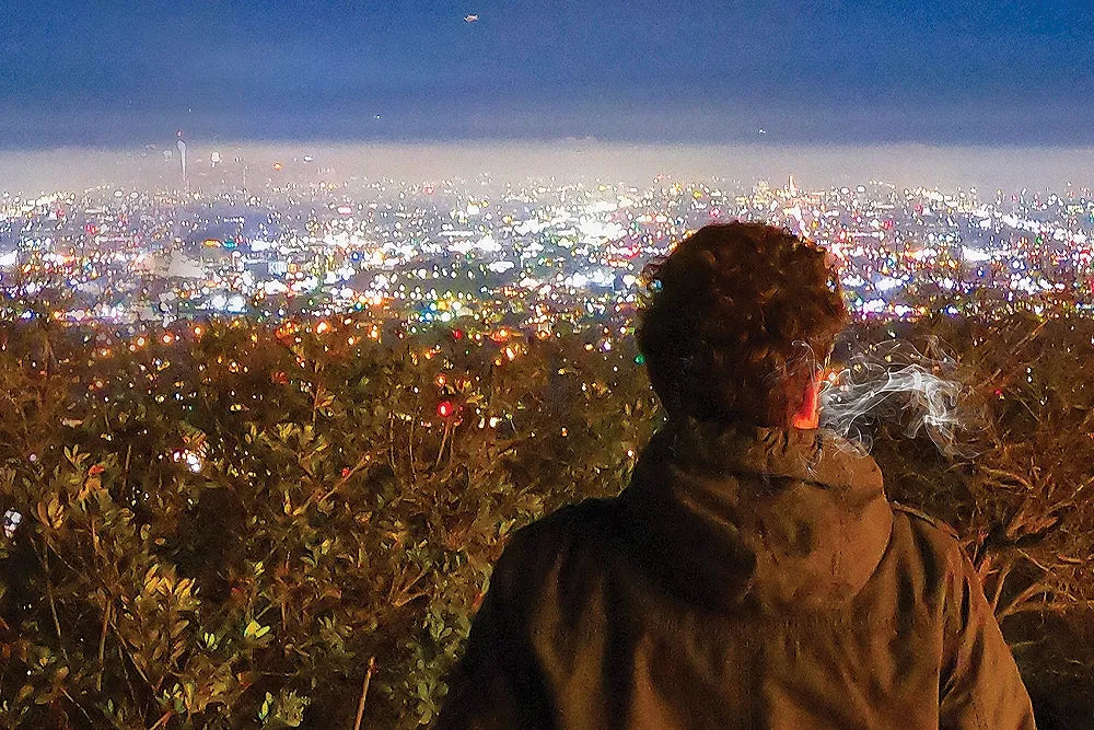 A smoker looks across the illuminated Los Angeles skyline at night.