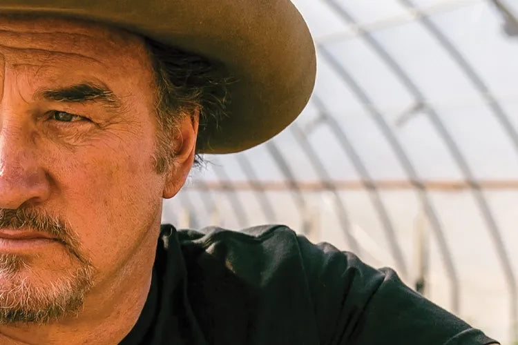 A close crop of Jim Belushi in a cowboy hat inside a greenhouse.