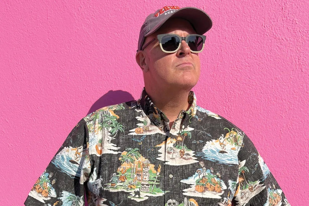 Adam Tschorn in a patterned shirt, cap and sunglasses against a pink wall.