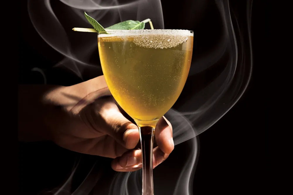 A hand holds a smoking green cocktail with a salted rim.