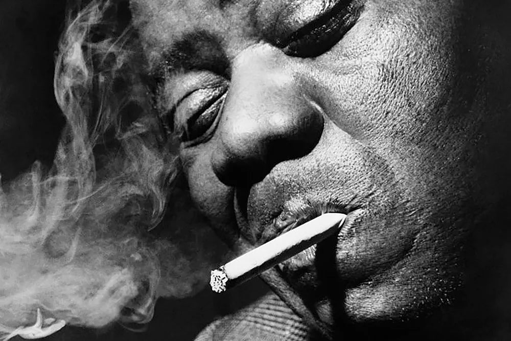 Black-and-white close-up of Louis Armstrong with a smoking joint at his lips.